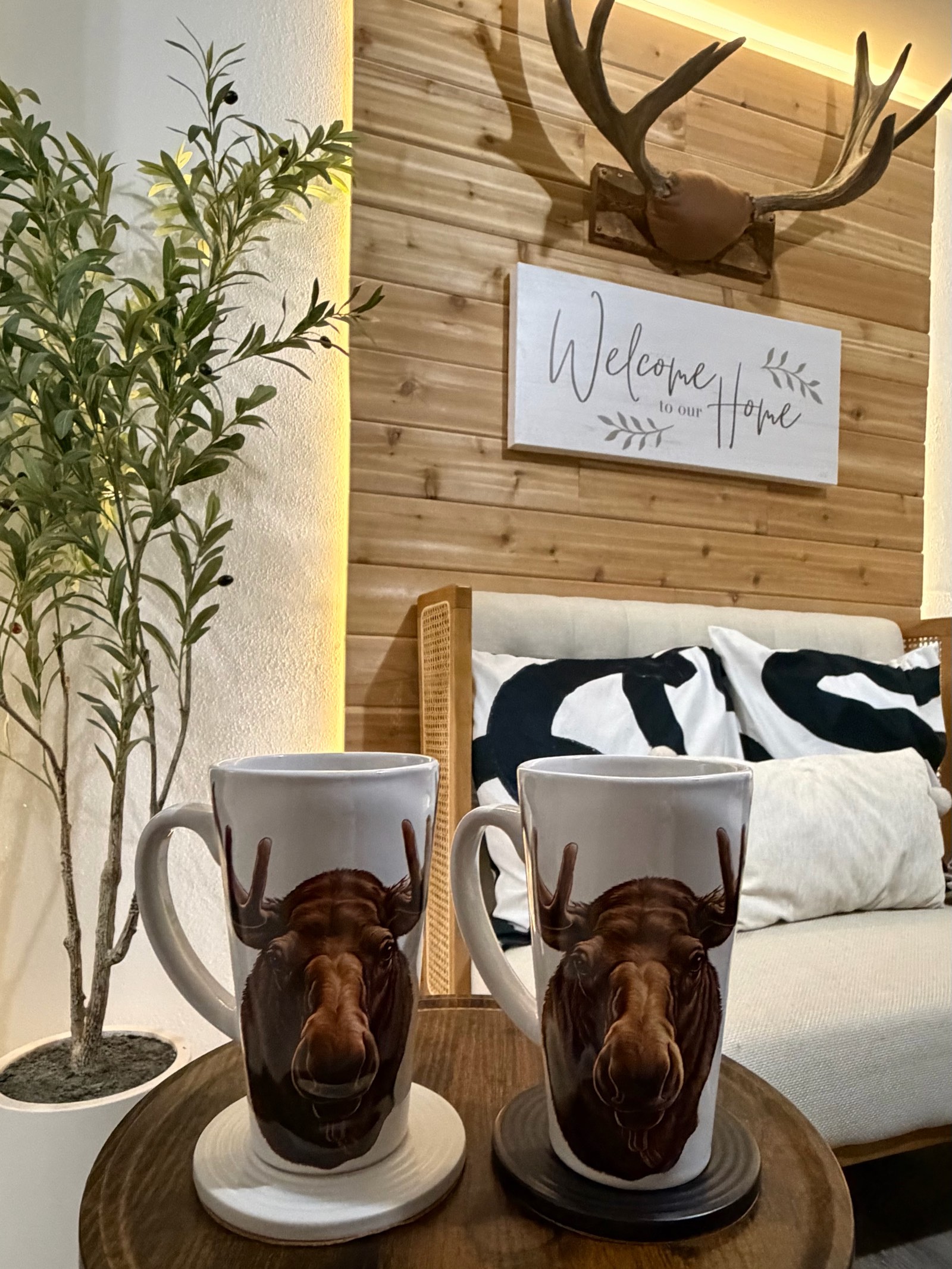 Cedar plank accent wall with a mounted set of antlers above a Welcome to our Home sign, a cream bench with black and white pillows, and two moose mugs on a side table.