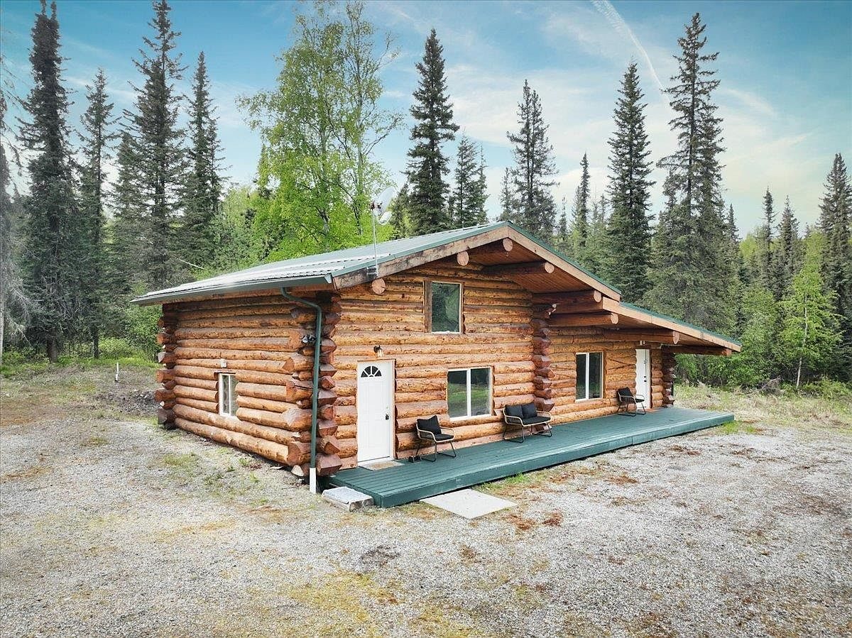 Front of the log cabin on Rambling Road with a green metal roof and a covered porch, surrounded by spruce and birch.