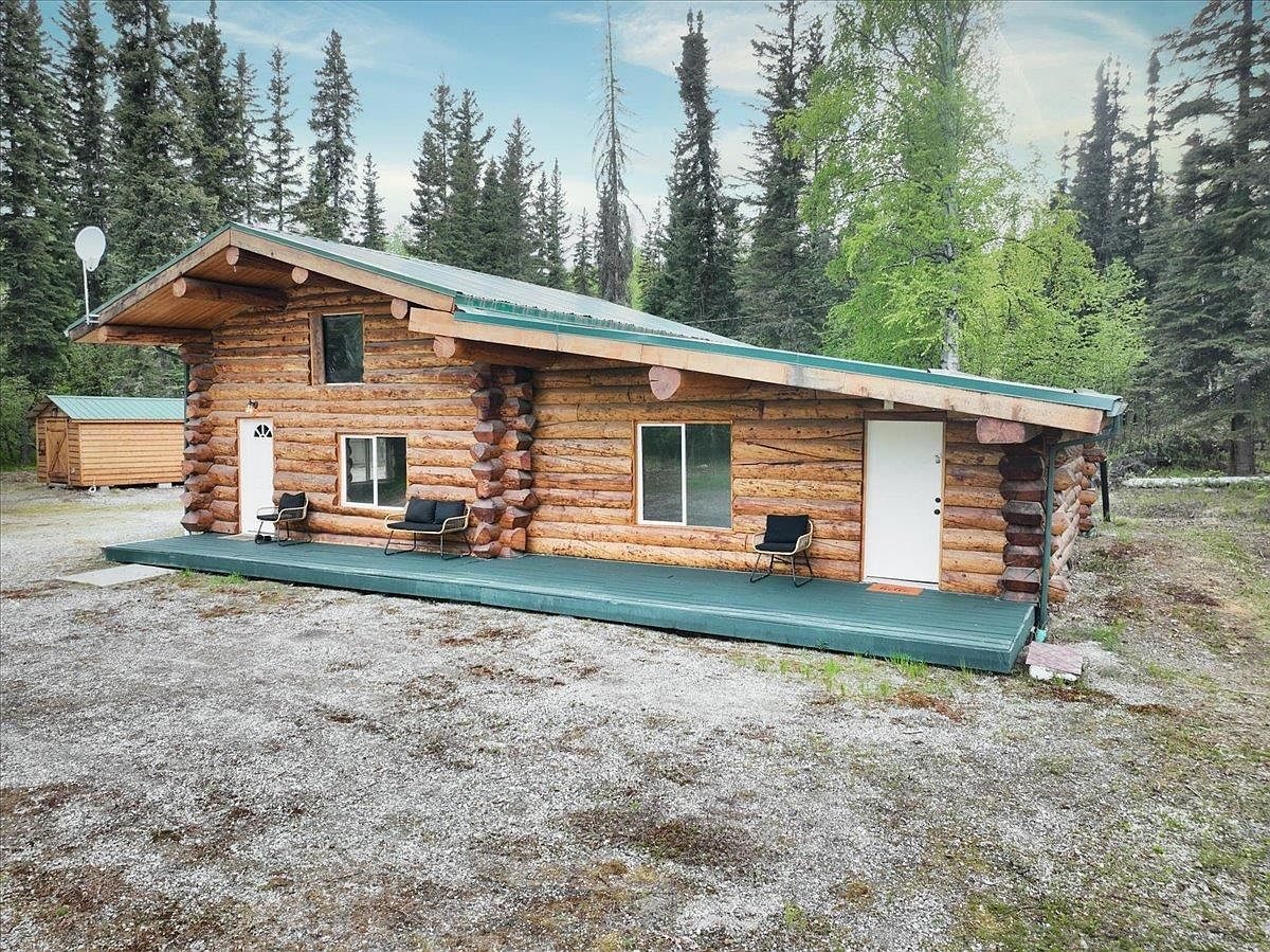 Side of the log cabin with its covered porch and a cedar-sided outbuilding in the trees behind.
