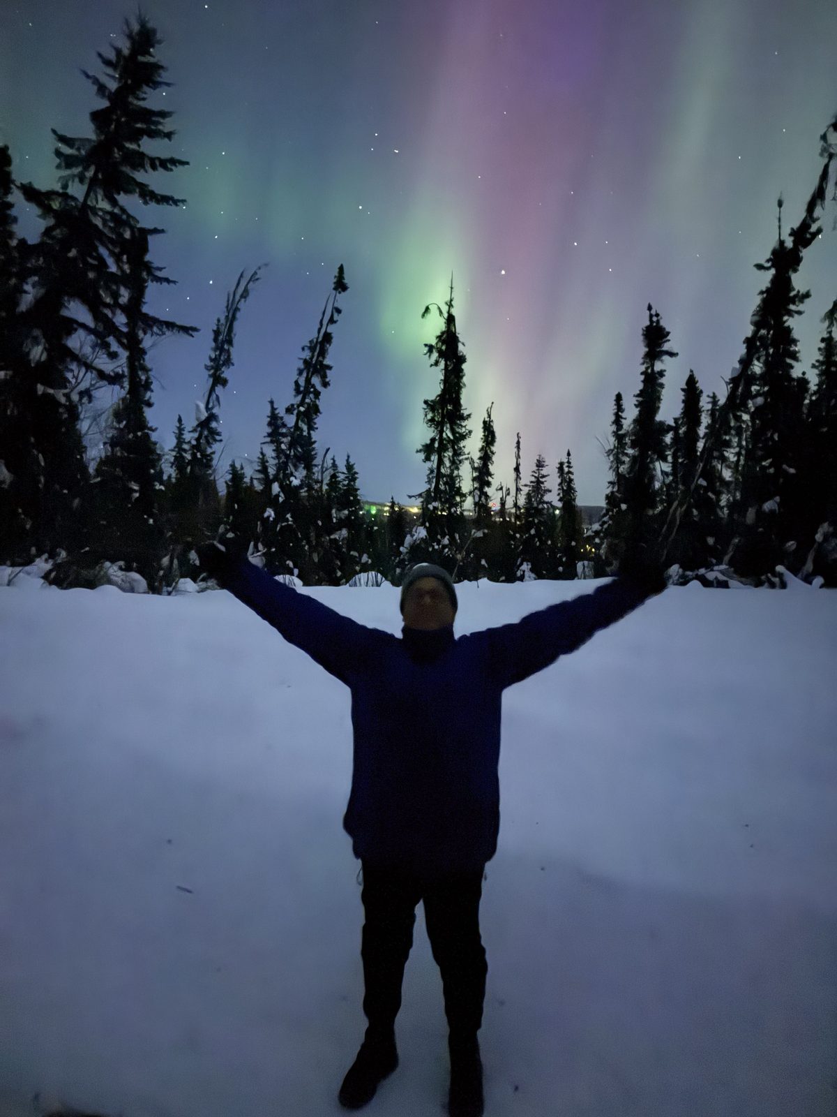Valdir standing in the snow with his arms raised under green and purple northern lights, spruce trees all around.